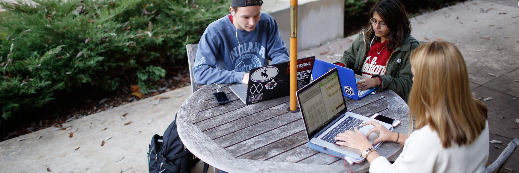 Three students listen to music and work on their laptops at an outdoor table on campus.